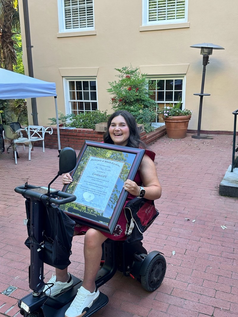 Ava, Kevin's daughter, smiling and holding her framed CarolinaLIFE graduation certificate from the University of South Carolina